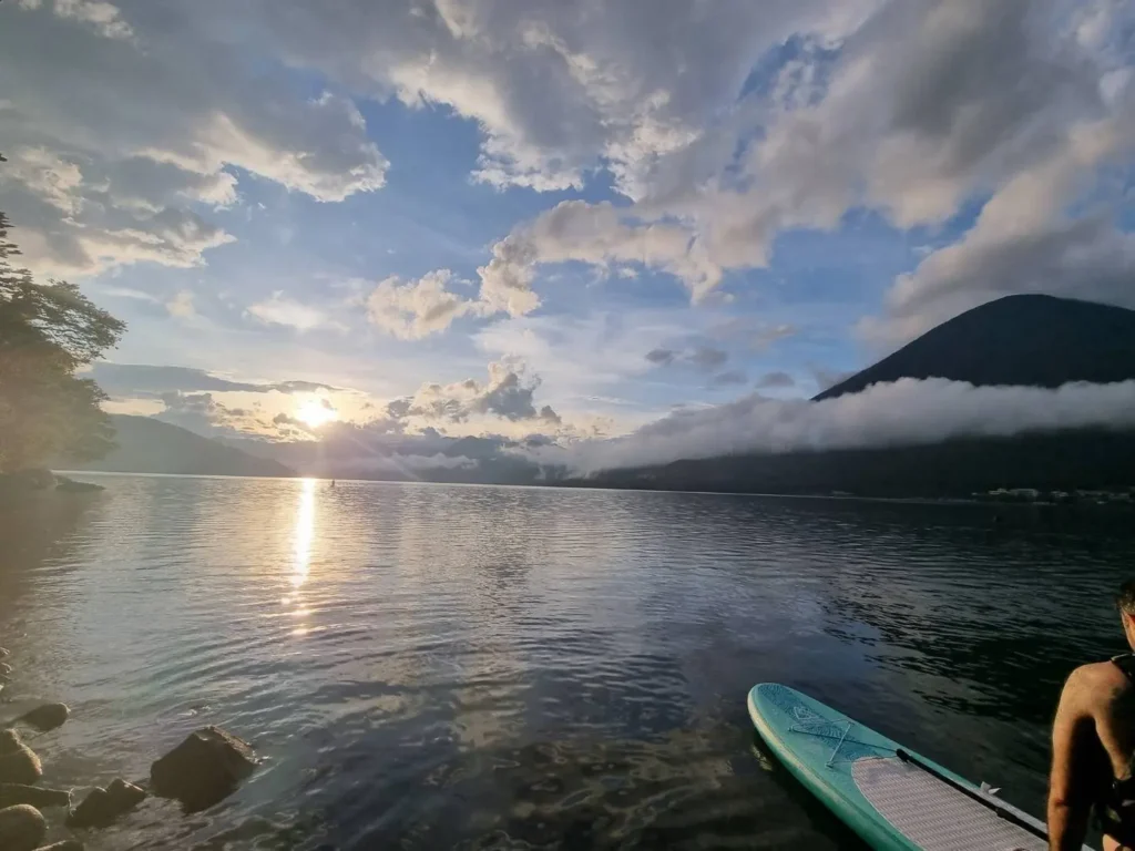 Man sitting on a stand-up paddleboard by the shore facing a lake at sunset