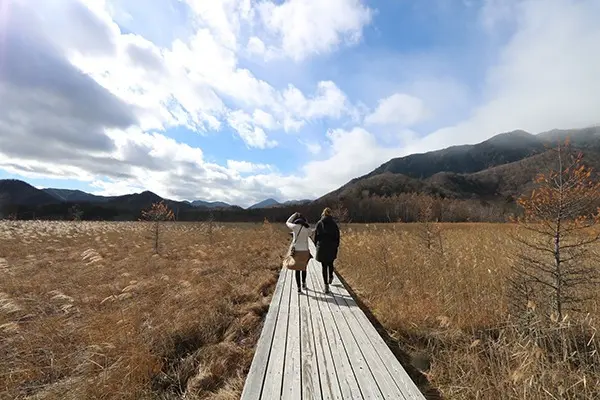 Two women walking on a wooden boardwalk through a field with mountains in the background