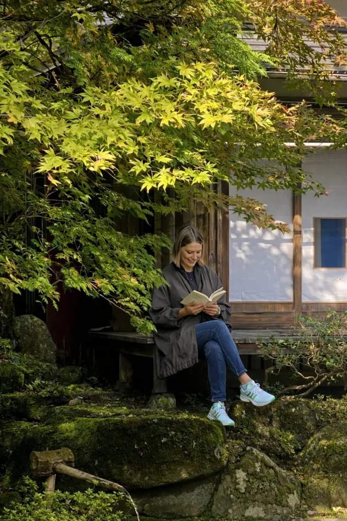 Woman reading a book while sitting on the porch of a Japanese house with rocks and tree leaves nearby