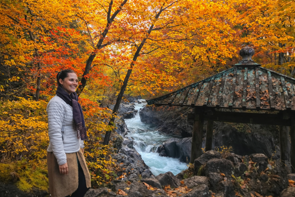 Woman standing beside a small stream near a roofed structure among trees